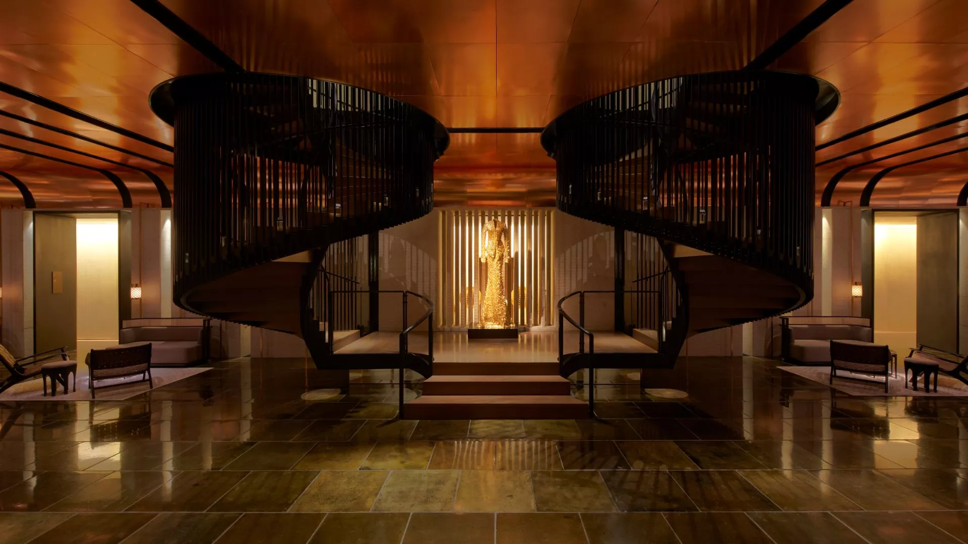 Elegant interior with twin spiral staircases flanking a golden sculpture and warm lighting at The RuMa Hotel and Residences